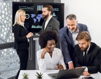 Positive purposeful multiracial business colleagues, working together over joint business project. Focus on mature blond woman and bearded man behind, using interactive screen in modern meeting room