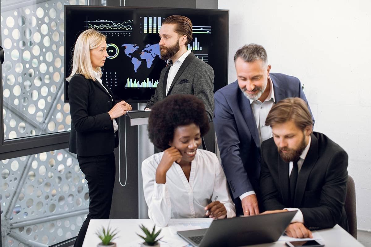 Positive purposeful multiracial business colleagues, working together over joint business project. Focus on mature blond woman and bearded man behind, using interactive screen in modern meeting room